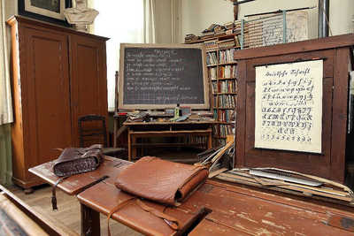 Blick in das historische Klassenzimmer mit Pult, Tafel und Schulbänken.